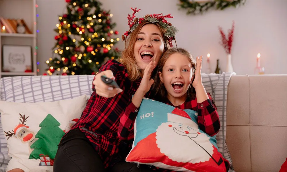 Fröhliche Mutter und Tochter sitzen in weihnachtlicher Atmosphäre mit der Fernbedienung auf dem Sofa